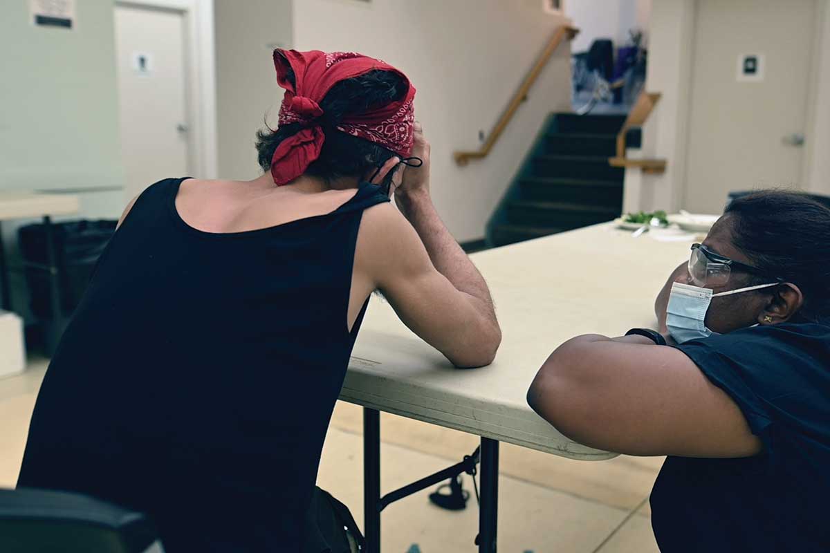 Woman with her back to the camera holding her head in her hands while another woman crouches down to talk to her