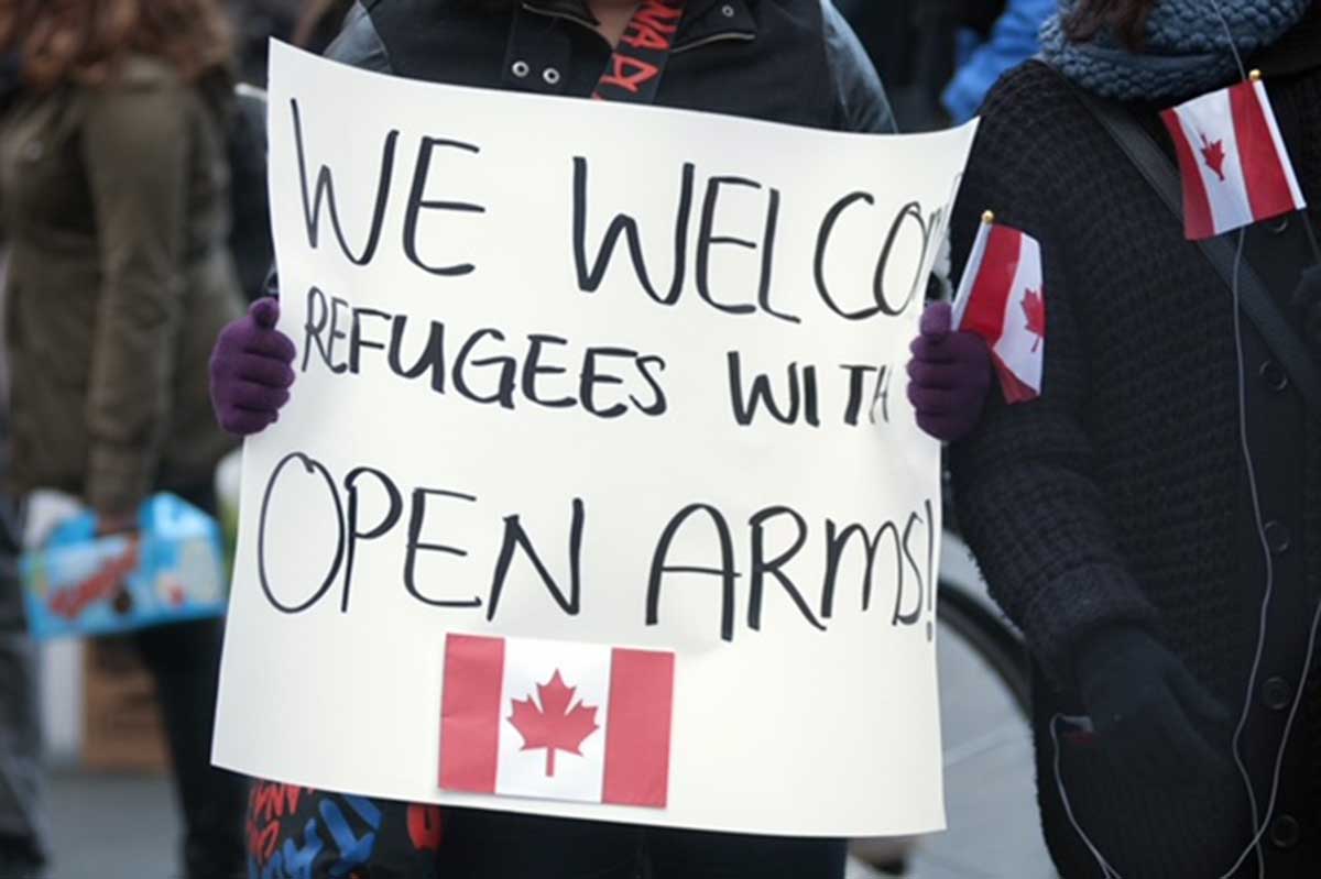Person holding a sign that says "We Welcome Refugees With Open Arms" along with an image of the Flag of Canada