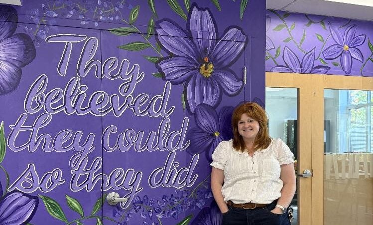 Anne Marie Hopkine stands beside a purple mural that says "They believed they could so they did."