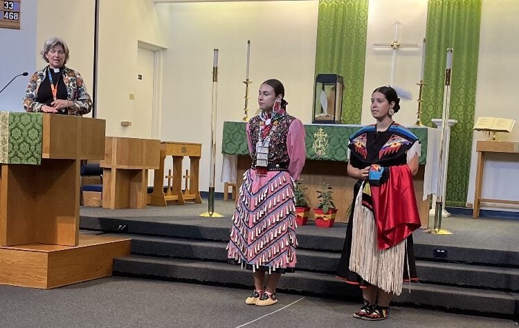 Priest introduces two young Indigenous women wearing traditional dance regalia.