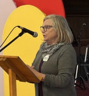 Shauna-marie Young speaks at a podium at the front of St. Albans church with Belong Ottawa's colourful logo in the background