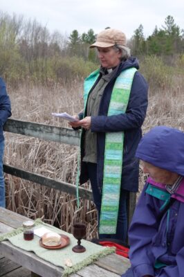 Dressed warmly for a chilly evening, a woman leads a Eucharist service on a wooden platform overlooking a pond.