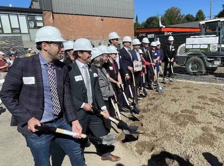 A large group of officials wearing white hard hats ceremonially dig shovels into the ground.