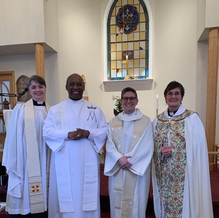 Four clergy members robed in white pose for a photo in front of a stained glass window.