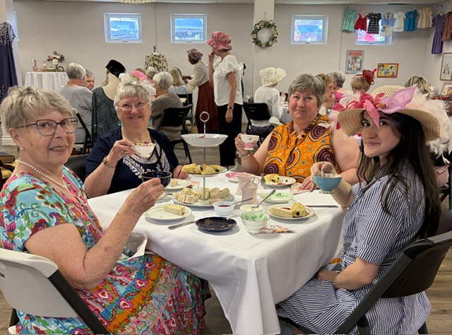 Women gather for tea, one wearing old fashioned hat.