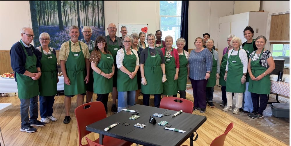 A large team of volunteers in green aprons pose for a group photo in the parish hall.
