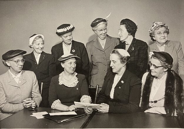 Black and white archival photo of a group of women in 1950s attire and hats talking at a table and posing for this formal photo.