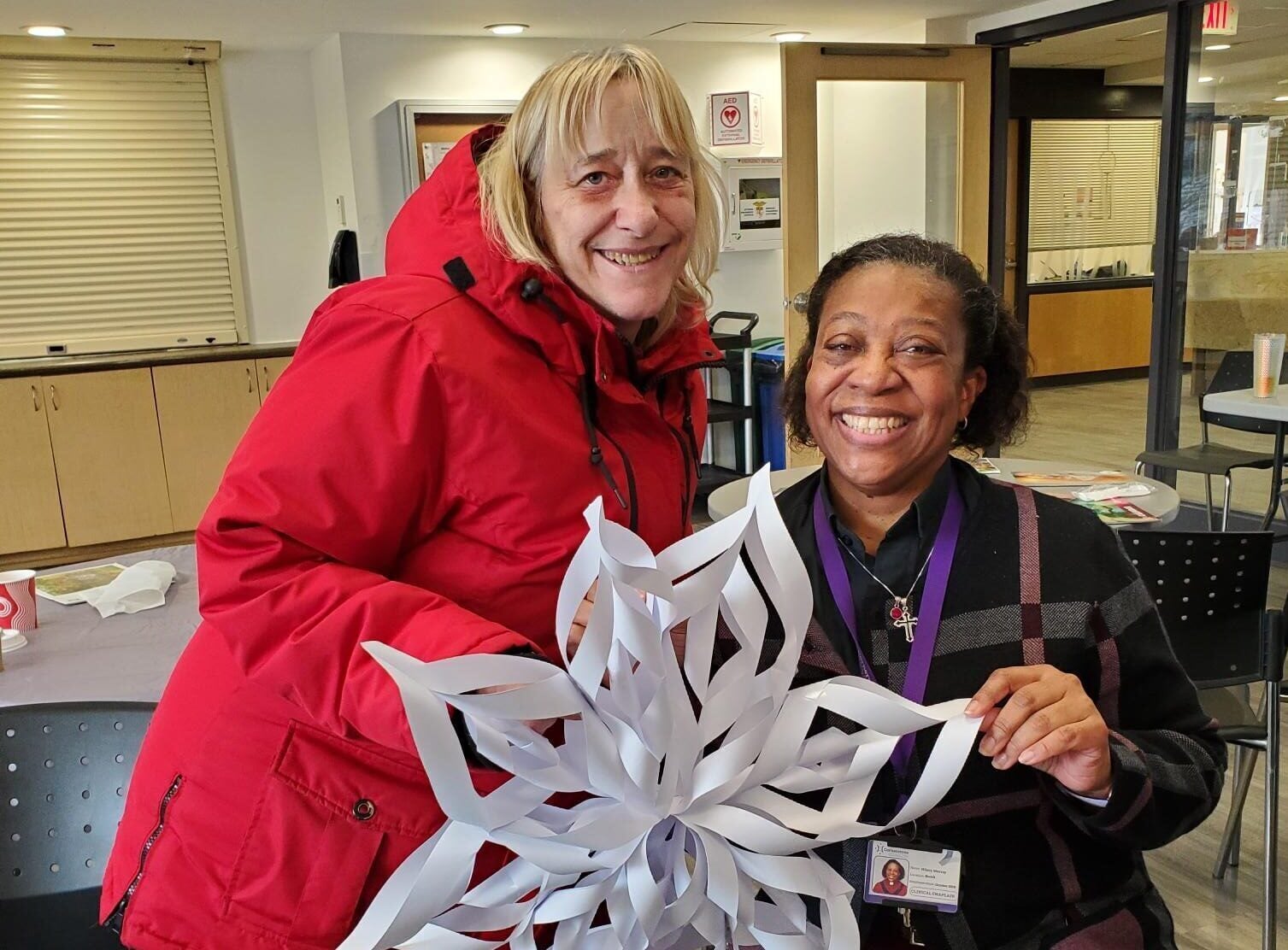 A woman in a red coat stands beside a sitting woman. Both are holding a decorative paper snowflake and smiling