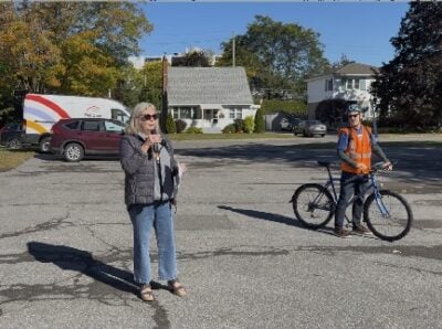 A woman with a microphone in a parking lot with a cyclist behind her
